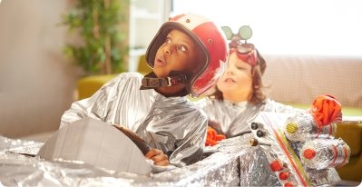 Two children in homemade silver astronaut suits sit in a DIY silver car spaceship, using their imagination to pretend they're flying through space.