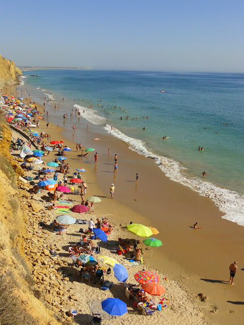 Estrecha y alargada playa llena de gente con el mar a su frente y acantilados a su espalda.