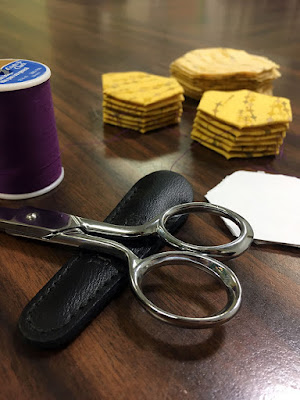 A close-up of a silver pair of scissors, the blades running out of frame to the left, resting on their black leather case, with a spool of plum thread and a small stack of white paper hexagons behind them, two small stacks of neat yellow hazeongs behind them, and fuzzily in the background, a rough-edged stack of yellow fabric hexagons, all on a dark wood table.