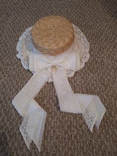 A straw hat covered in lace and ribbon. The brim of the hat is covered in lace. There is a bow at the back with the BTSSB charm hanging. The ribbons of the bow are very long and have a layer of lace and ribbon.