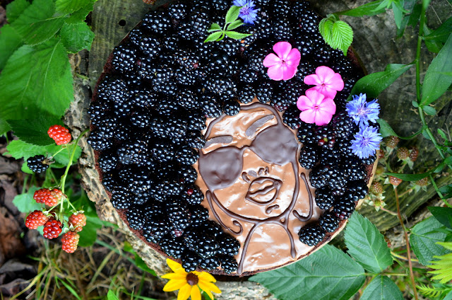 Bild von einem Schoko-Zucchini-Kuchen, der mit Brombeeren belegt und mit einem Gesicht aus Schokolade und frischen Blumen verziert ist.