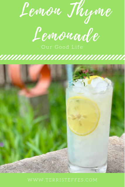 A glass of lemonade, with lemon slices and a lot of ice and a sprig of thyme in the glass sitting on a rock wall.