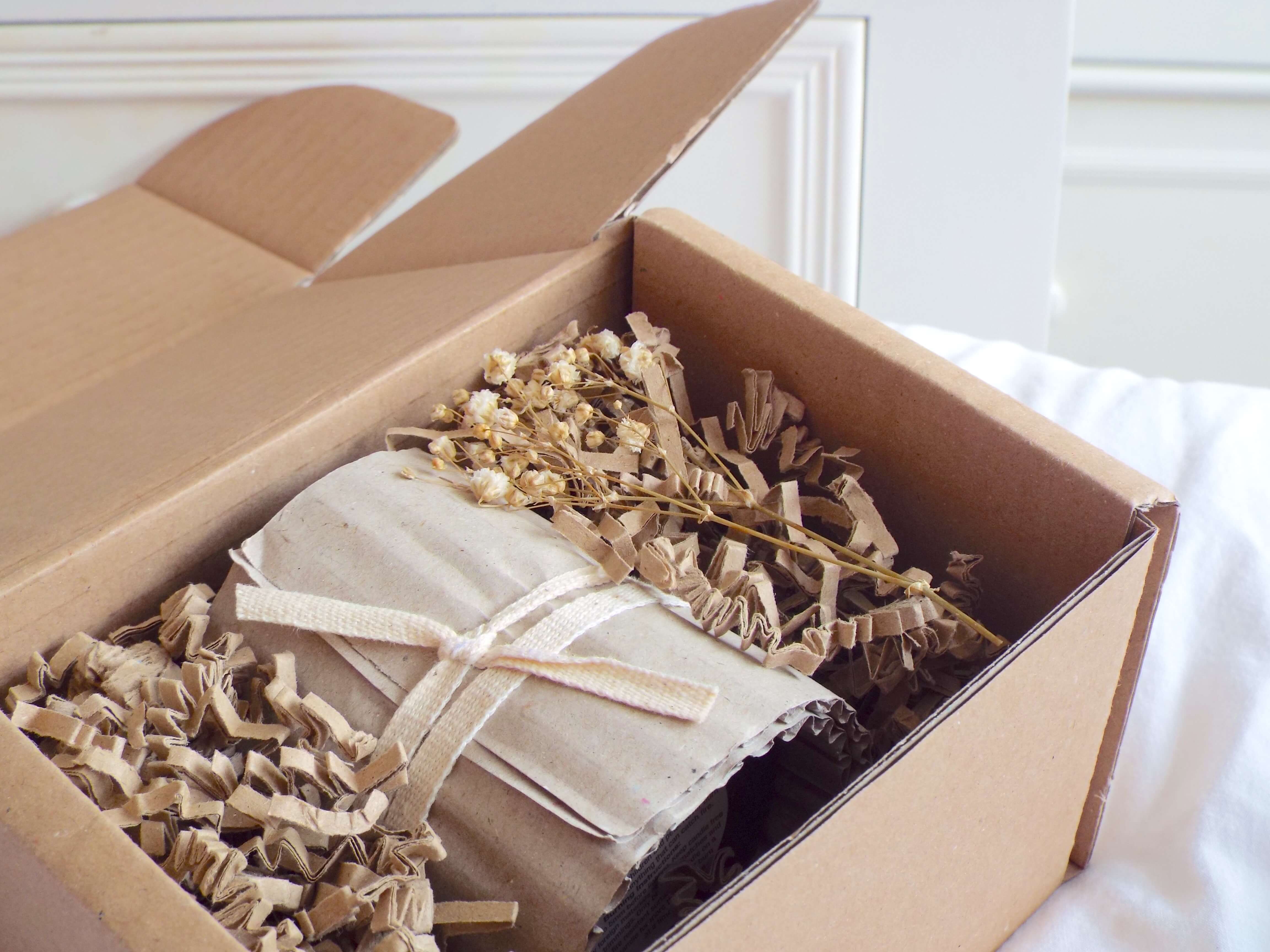 Landscape view of That Hygge Life Candle nestled amongst paper crinkle packing, on white bedding. a small, dried flower sits inside to the side of box.