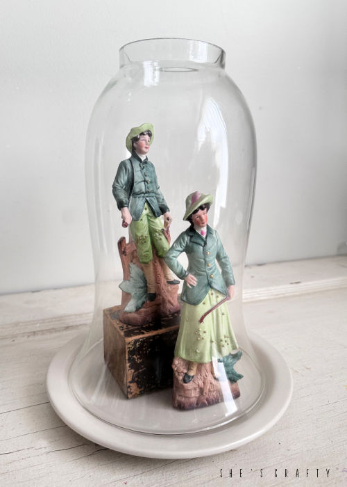 Man and woman figurines underneath a cloche.