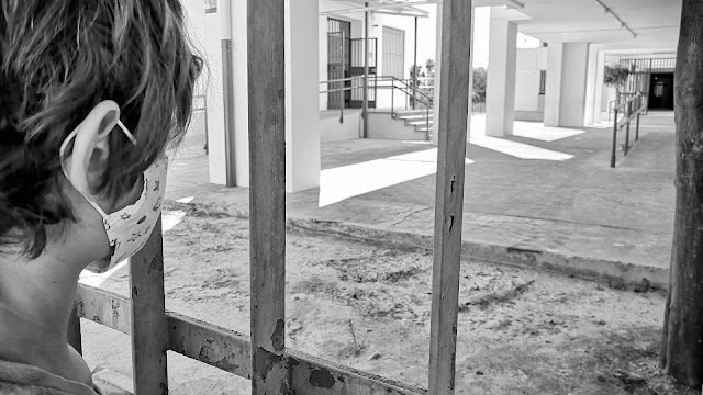 Javi, de espaldas con su mascarilla puesta, mirando al patio del cole a través de la reja de la entrada.