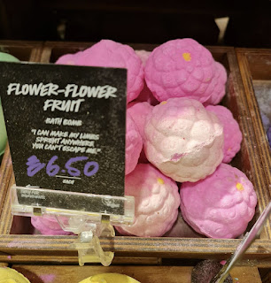 A light golden brown square box containing a cluster of oval light pink bath bombs with floral engravements in them next to a black card that says flower-flower fruit bath bomb in white font on a bright background