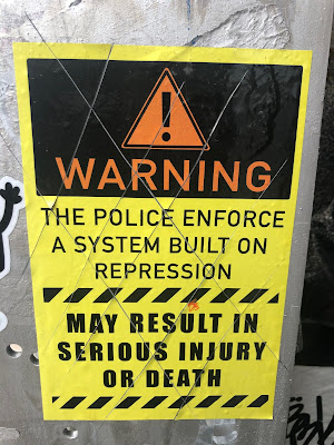 Sticker with "Warning" exclamation point symbol, says, "Warning: The police enforce a system built on represson, may result in serious injury or death"