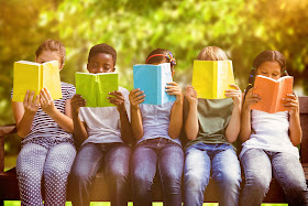 A row of children of different ethnicities sitting on a park bench, each reading a book