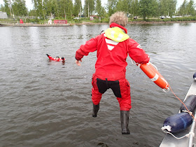 Henkilö hyppäämässä pelastautumispuvussa veteen pelastusrenkaan kanssa, taustalla huitoo toinen pelastautumispukuinen henkilö vedessä
