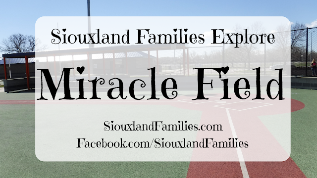 in background, a red and green rubber baseball field with white painted lines at Miracle Field in Riverside Park in Sioux City, Iowa. In foreground, the words "Siouxland Families Explore Miracle Field"