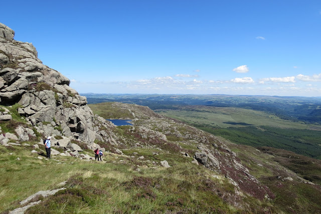 A view back down the path beside the rocky ridge - the lake lies below and beyond it, rolling hills into the distance.