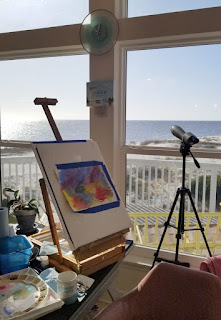 A photograph of an easel and camera set up next to a large set of windows overlooking a sparking blue ocean.