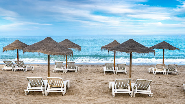 Playa con sombrillas y hamacas sobre la arena y el mar con oleaje al fondo.