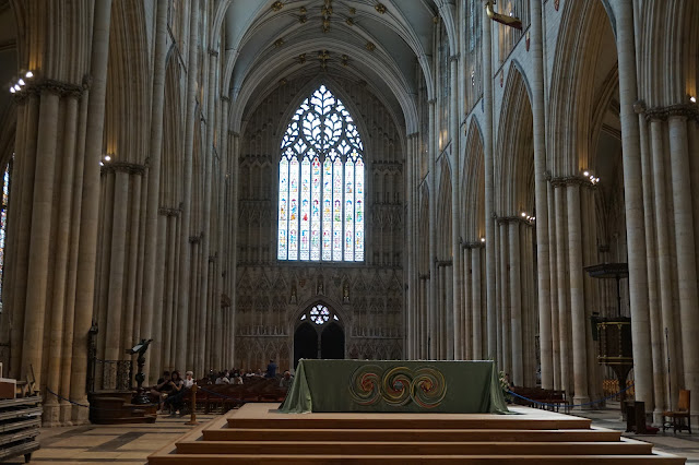 The nave of York Minster, with an alter on a stone platform with people sitting in seats, and a large stained glass window