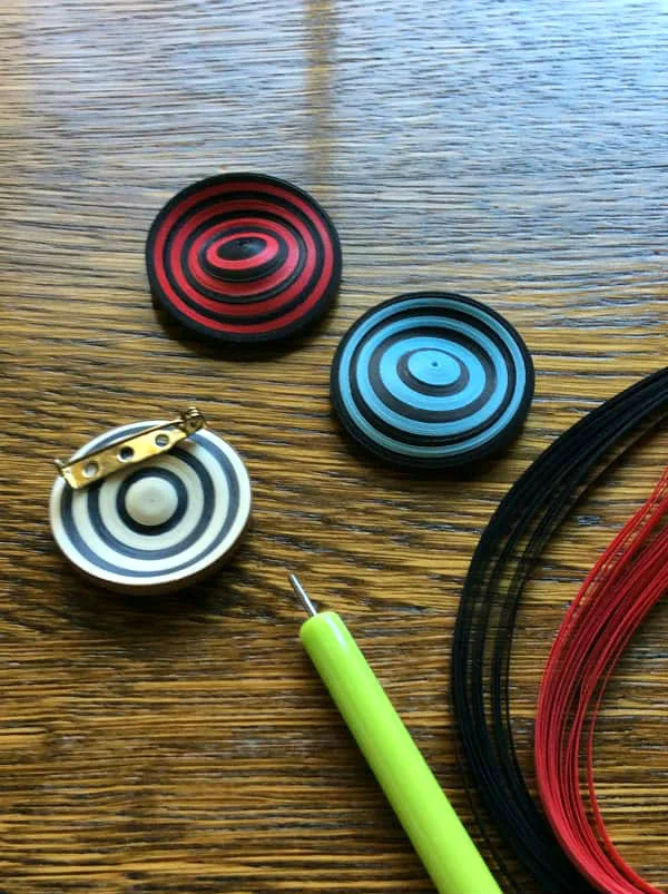 three circular, striped paper brooches on wooden surface next to quilling tool and red and black quilling strips