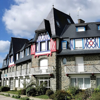 Photograph of a large building on the corner of a road; its first two floors are stone, with doors and shuttered windows. The upper floors are half-timbered, with timbers painted in bright colours, and a hipped mansard roof with several steeply-pitched gables.