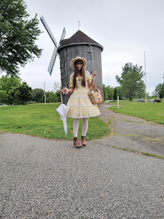 A lolita girl (me) standing in a park in front of a windmill. Yellow dress from Baby, the Stars Shine Bright, white socks, brown boots, a straw hat and holding a straw bag in one hand and a white parasol in the other.