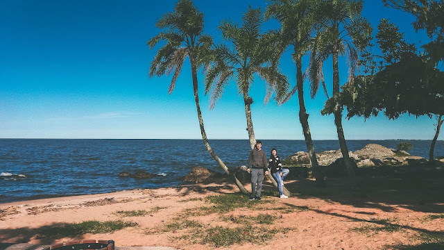 Casal encostado em árvores, na beira da praia