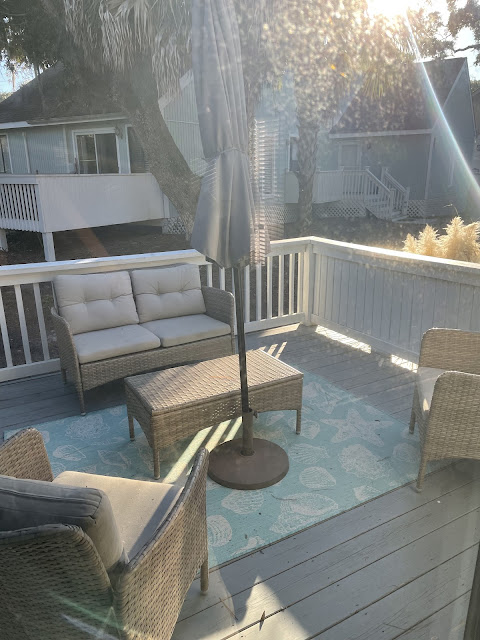 A view of the porch at the house we rented. The porch is painted light gray. It has two outdoor armchairs and a loveseat all with cushions. There is an outdoor coffee table in front of the loveseat with an outdoor umbrella that is currently closed. The umbrella is gray in color as are the cushions. The furniture is made of wicker material and is a brownish gray. There is an outdoor area rug which is also gray and has seashell designs on it.