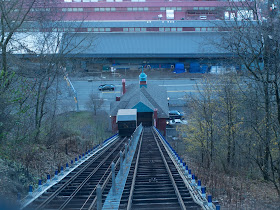 A view looking down an incline with tracks for cars, passing through trees and up to a red building on a two-line road with large buildings on the opposite side.