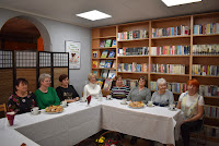 Zdjęcie przedstawia siedzące przy stolikach panie, które przybyły na spotkanie z okazji Dnia Kobiet. Na stolikach kawa, cherbata, słodki poczęstunek oraz kwiaty.