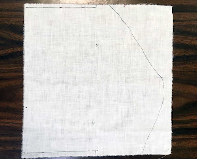 A square of white linen with frayed edges on a dark wood table, with a rough right-pointing curve marked on it in black pen.