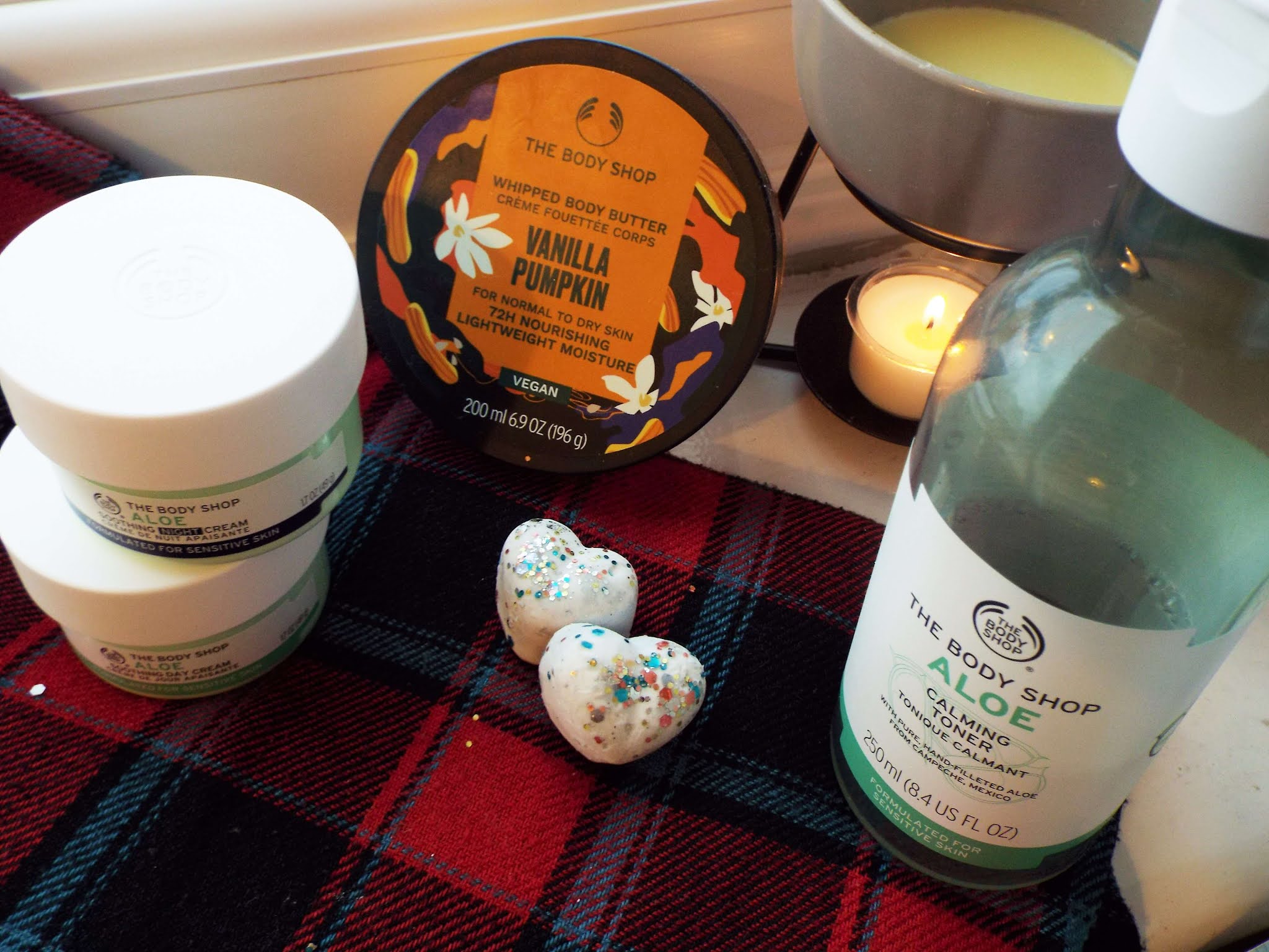 Cluster of October favourite beauty products, sat on tartan scarf in nook of windowsill, with wax melter burning away in the background.