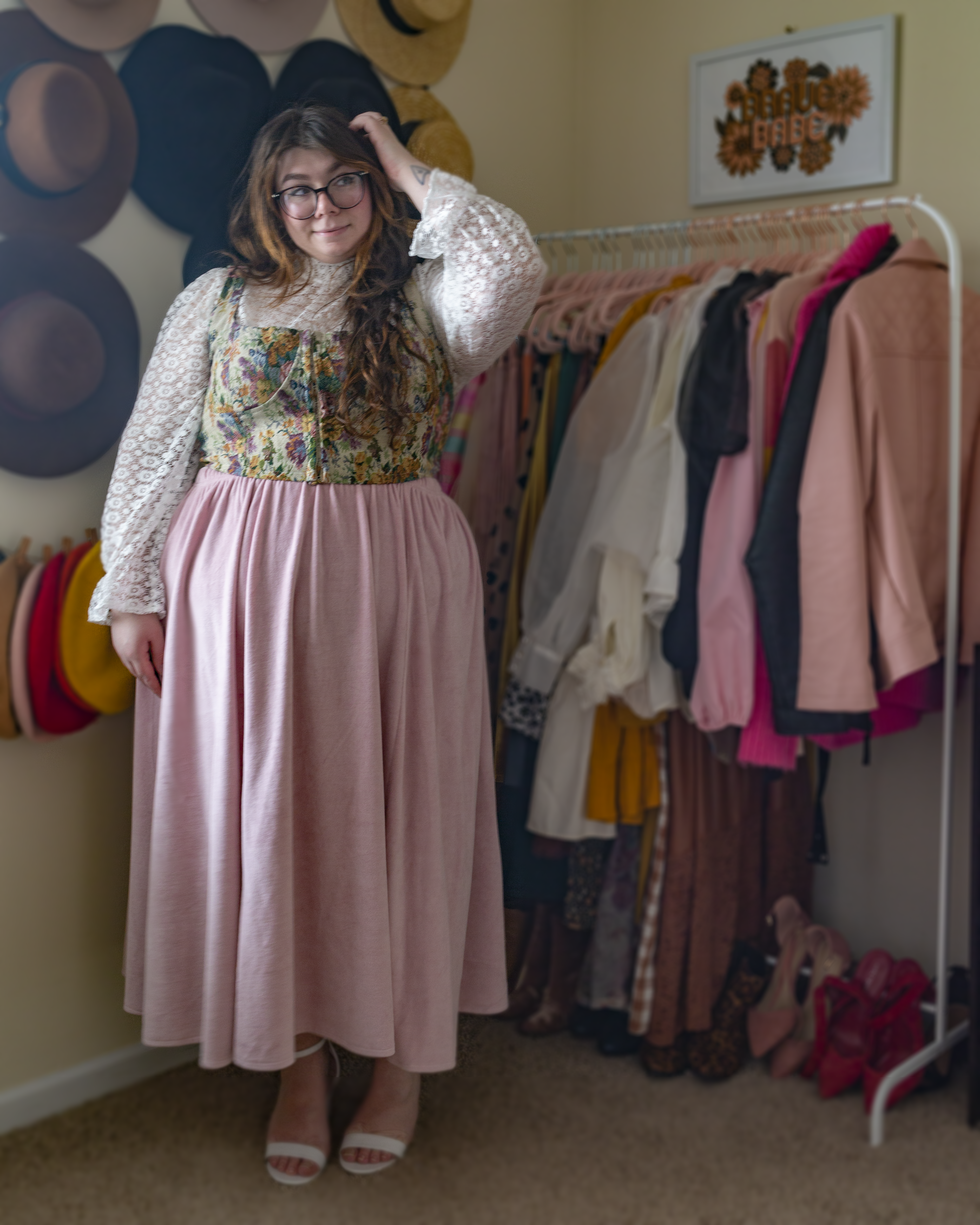 An outfit consisting of a white floral lace top with long sleeves with a ruffle hem, with a pink, green and yellow floral corset and pink velour midi skirt and white ankle strap heels.