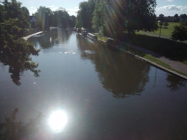 Photograph of the River Cam on a sunny, summer day, from one of the many bridges. There are rainbowed lens flare strands coming from above, but the sun is only seen reflected almost painfully brightly in the broad, tranquil river, on which there are swans (white scribbles on the water that might also be other stuff!). Large trees in full leaf frame the river and rowing club buildings are visible on the left-hand side, with canal boats moored on the opposite bank. The patch of common land seen on the right is a brilliant green, and there are a few fluffy clouds in the light blue sky.