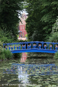 Yellow water lilies in front of a bright- to dark blue iron bridge under large trees with a red painted castle in the distance.