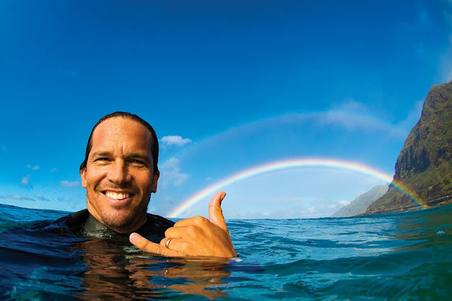 Clark Little fazendo hang loose no mar. Atrás dele uma costa rochosa e um arco-íris.