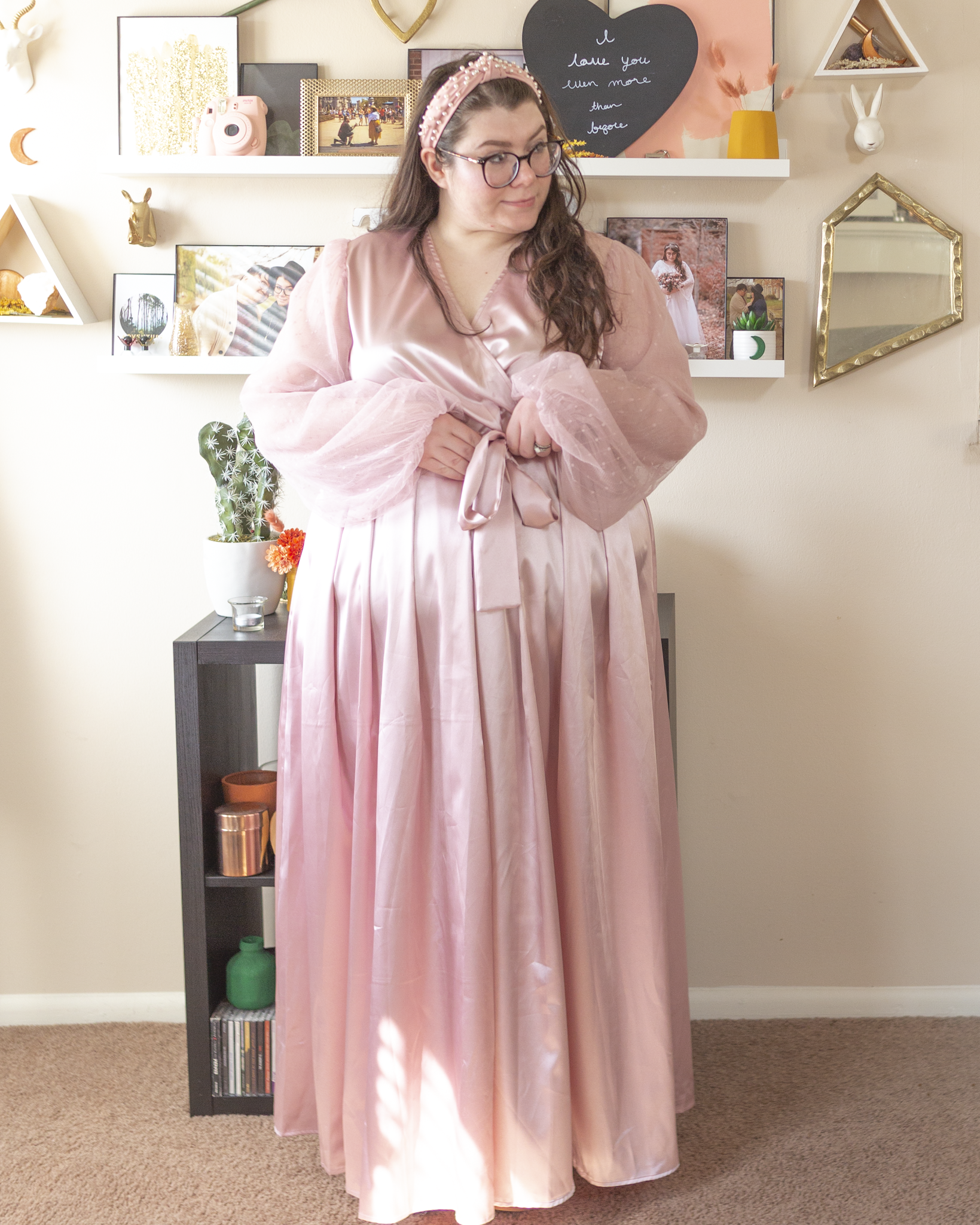 An outfit consisting of a pearl encrusted pastel pink headband, a pastel pink satin wrap dress with sheer bishop sleeves.