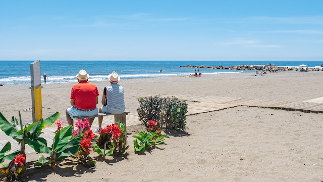 Dos personas sentadas de espalda en la zuna de las duchas de una playa de arena con las azules aguas del mar al fondo.