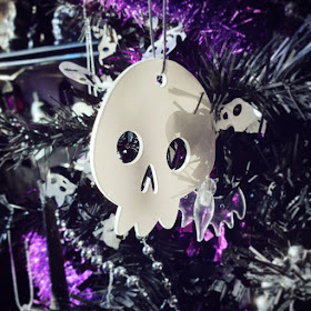 A cartoonish white acrylic skull, very glossy, hanging in the black Christmas tree, with a clear bat-shaped LED string light, silver bead garland, purple tinsel, and black tinsel with white skulls. The shelve and lantern-styled string lights are visible in the background, out of focus, in the upper left 