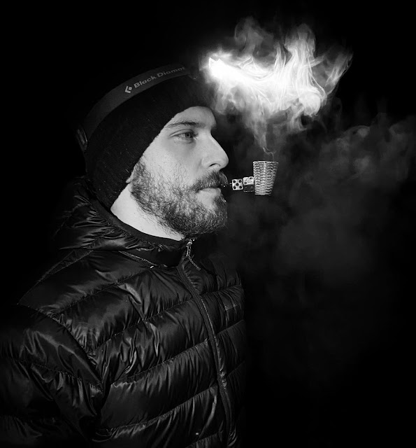 Wearing shiny puffy fleece and a hat standing from side view blowing out smoke from a pipe and mouth