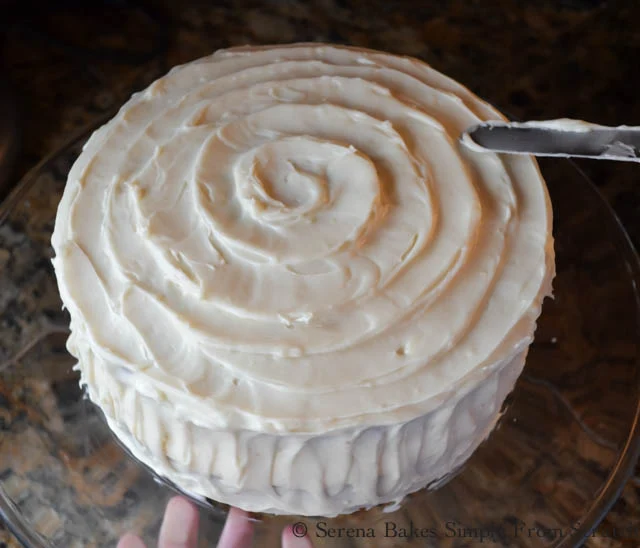 Cream Cheese Frosting made into a large swirl on the top of Carrot Cake on a glass cake stand.