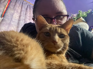 In the foreground is a ginger cat in a loaf position with his head turned to look over his right shoulder to look at the camera. Behind him is a brunette, bespectacled, middle aged white woman, wearing a black T-shirt, leaning forward to kiss him on top of his head. They're both looking at the camera, and the cat's left eye is slightly cloudy.