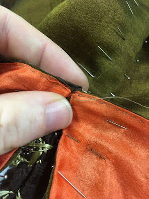 A close-up of a white hand pinching the intersection between two panels of fabric, sheer peach on the near side and green-gold on the far side, with pins anchoring the folded-in edges of all pieces.