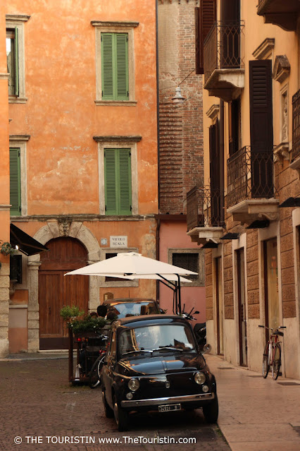 A little black old Fiat 500 model parks next to a cafe with white sun umbrellas on a cobbled street lined by four-storey orange-painted period houses with closed green wooden shutters.