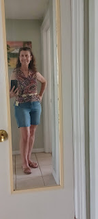 Woman wears a colorful paisley shirt that is sleeveless and zips up the front. A carved belt is over the shirt. She also wears denim shorts and light brown sandals.