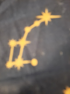 Photo of yellow print constellation with yellow line joining representations of stars . The figure looks like a  square with a line twice its height  extending on a 20 degree angle from the top left corneŕ where there is another large star represented that looks a bit like a peafowl head a short line an eighth the height of the box to the right and ending in a smal dot looks like a beak