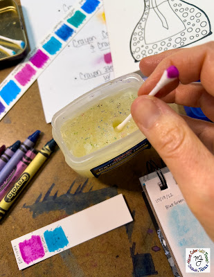 A clean cotton swab is being dipped into an open tub of petroleum jelly that is obviously used only for art projects as it has specks of color floating in the jelly.