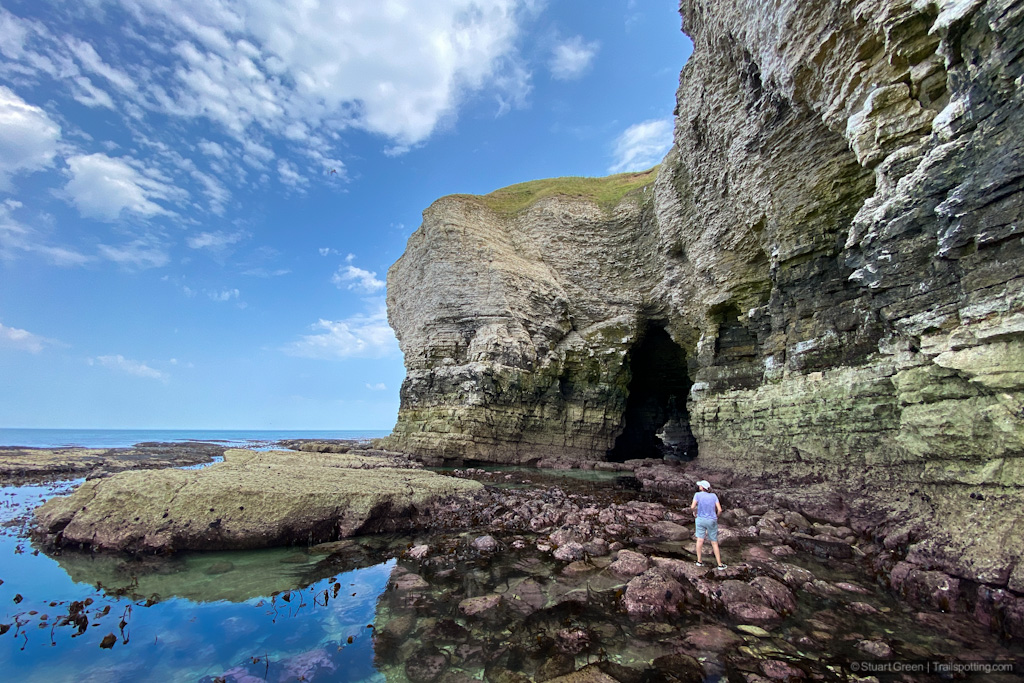 Visitor stands on purple seaweed covered rocks, with cliffs high above and more sea caves in the distance.