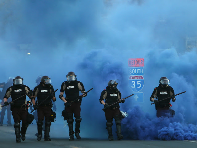 Minneapolis police in riot gear advance in a line through billowing blue tear gas smoke, with their batons out.