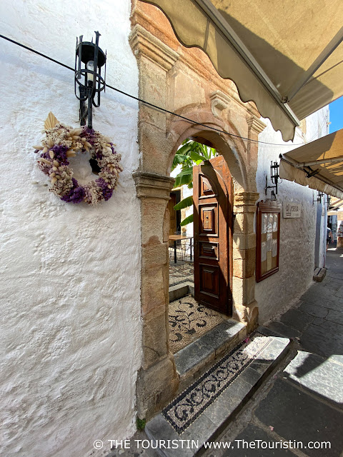 A white and purple wreath next to an opened wooden door on a with pebbles tield lane that is lined by whitewashed houses.