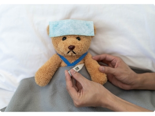 A teddy bear laying on a bed, looking sad and ill with a wet towel on its head, while getting its temperature checked.
