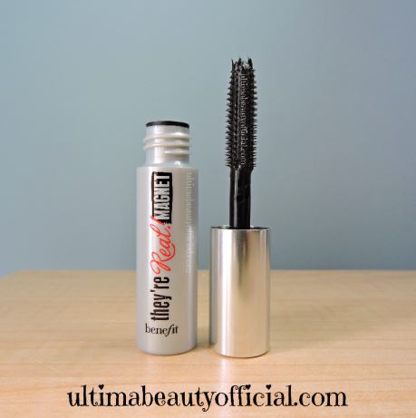 Opened grey mini mascara tube with mascara wand next to it. Printed on Tube: Benefit They're Real! Magnet