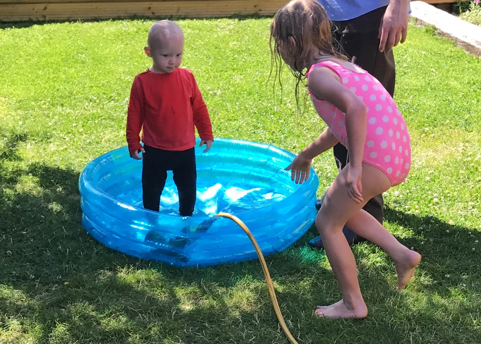 Filling a small paddling pool with a hose while a toddler stands in it fully clothed