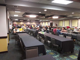 A view of the free play area. A hall in the hotel with a number of long tables surrounded by chairs. Many of these chairs are occupied by people playing a variety of board games.