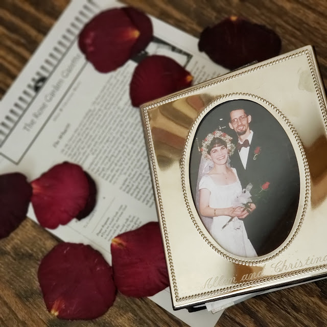 A small silver photo album with a photo of bride and groom and the words "Allen and Christina" on the front. Beneath that some hardwood floor is showing around the edges, with a printed newsletter called "The Rose Garden Gazette" and scattered red rose petals